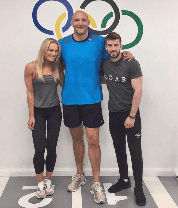 Ben Kay standing between Sarah Lindsay and a ROAR trainer in front of an Olympic rings mural at ROAR Fitness London