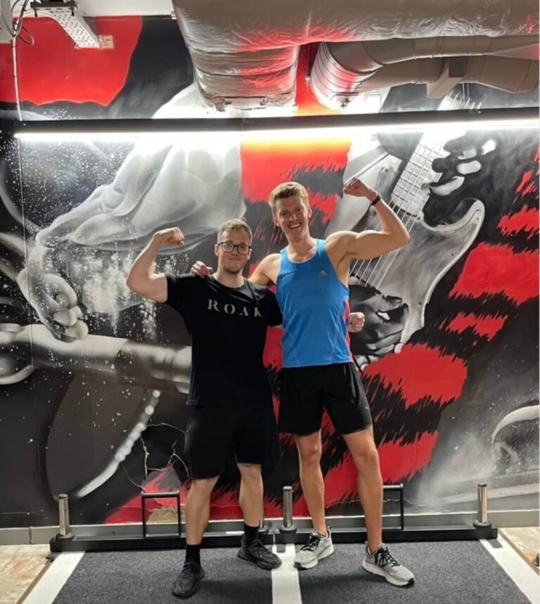 Chris Baber and his ROAR personal trainer flexing in front of the gym mural at ROAR Fitness Kensington