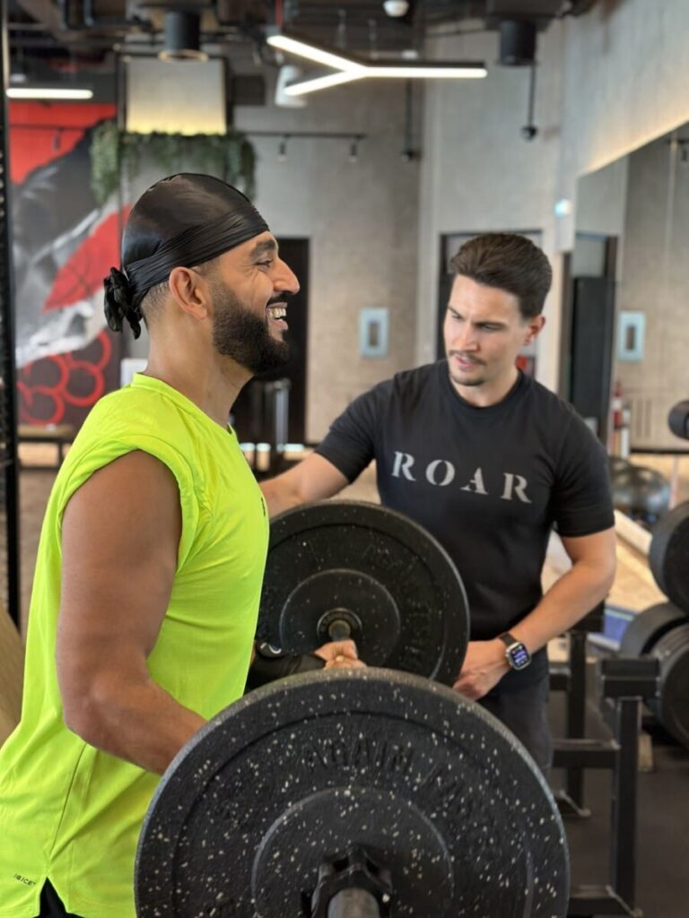 DJ Bliss smiling mid-set with a barbell as a ROAR personal trainer supports his shoulder at ROAR Fitness Dubai