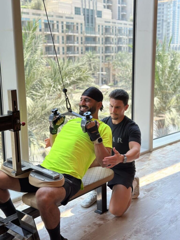 DJ Bliss using a cable machine while a ROAR personal trainer coaches him, with the Dubai skyline behind
