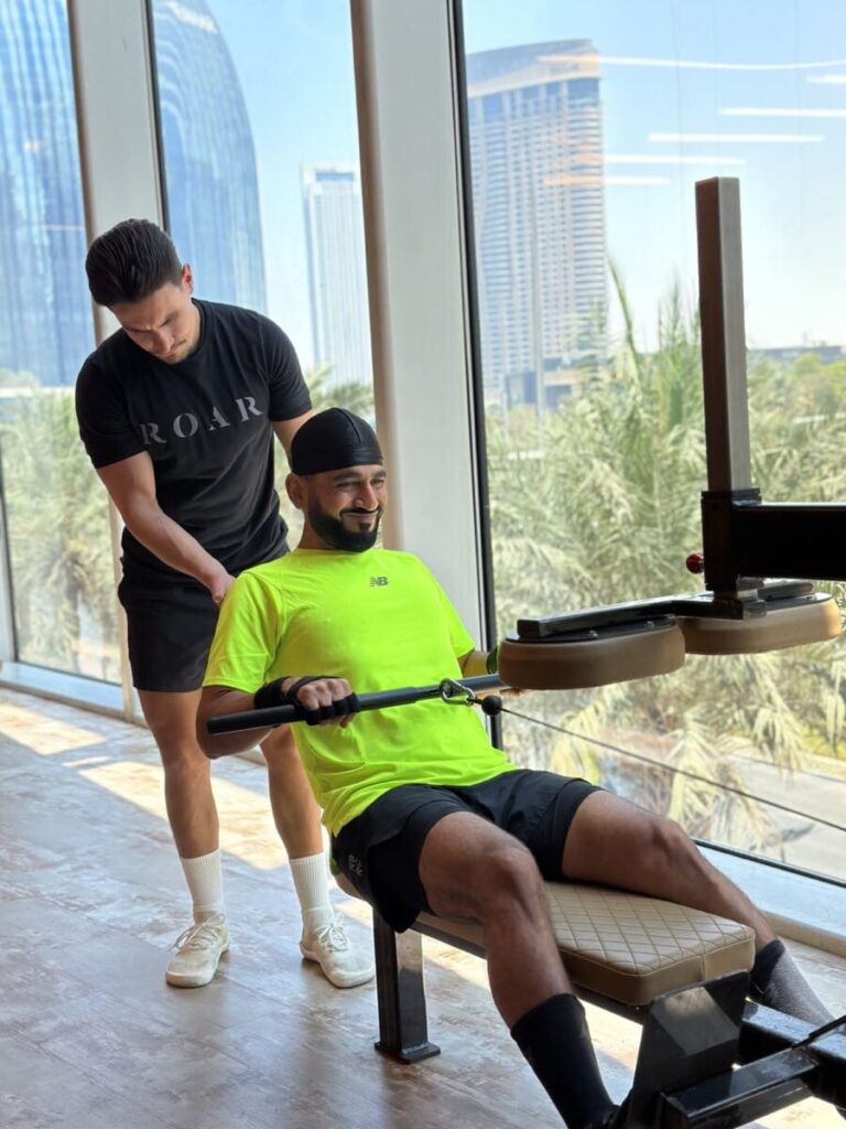 DJ Bliss seated on a leg machine with a ROAR personal trainer behind him at ROAR Fitness Dubai