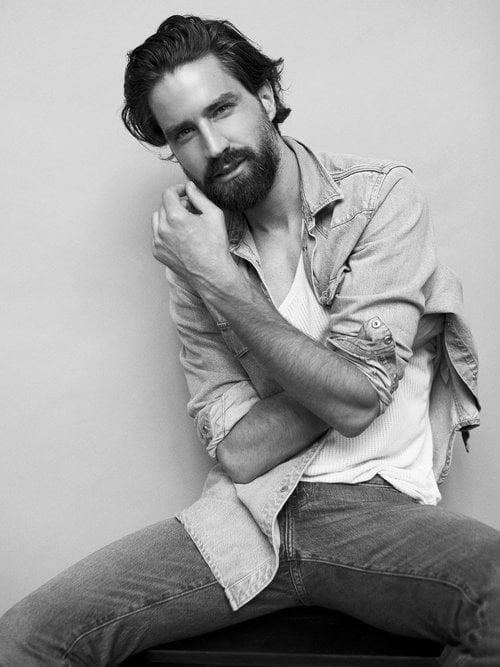 Black and white studio portrait of Jack Guiness seated in a denim shirt