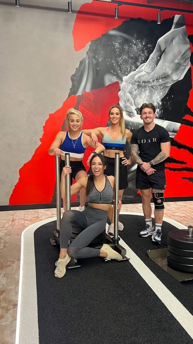 Aimee Fuller and Katya Jones posing around a sled on the training track with ROAR trainers at ROAR Fitness London