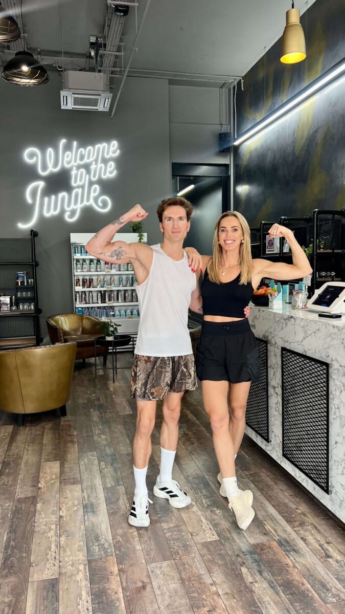 Oliver Proudlock flexing beside a ROAR trainer under a neon sign in the reception at ROAR Fitness London