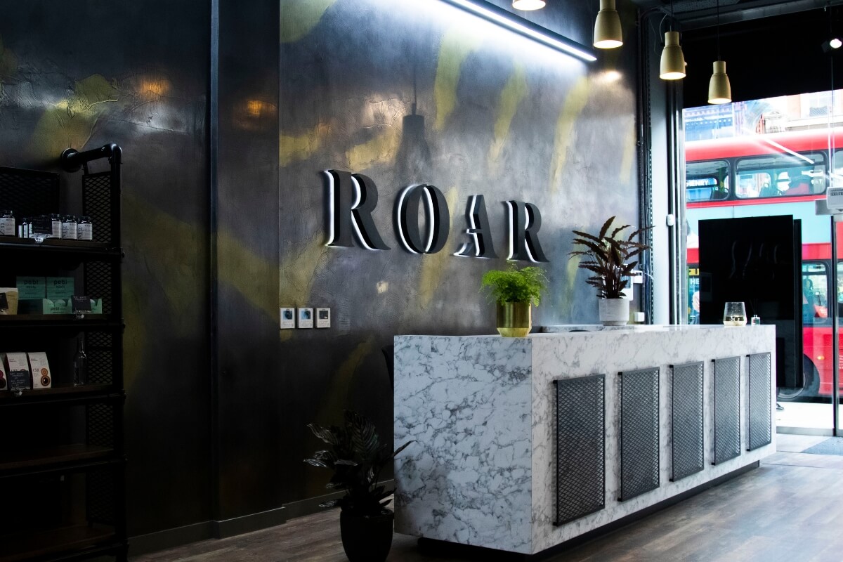 ROAR Fitness Kensington reception desk in marble and black mesh beneath a backlit ROAR sign on a dark textured wall